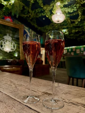 A close-up, eye-level shot of two glasses of rosé champagne placed on a wooden table, set against a dimly lit, atmospheric bar interior decorated with greenery and a chandelier.