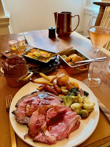 A high-quality, professional-grade photograph of a premium Sunday roast at The Connaught Grill. The image features a perfectly cooked beef rib, a large Yorkshire pudding, and seasonal vegetables, accompanied by a glass of rosé champagne. The lighting is soft and natural, highlighting the textures of the food, and the shallow depth of field creates a clean, focused composition.