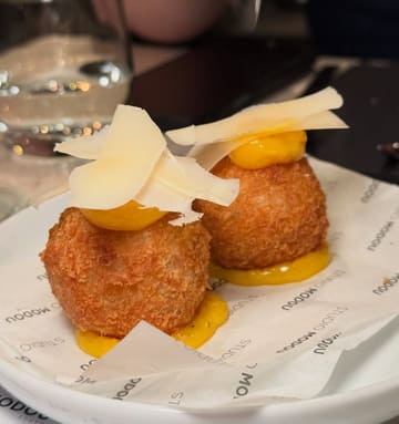 A close-up, eye-level shot of two Cacio e Pepe arancini balls, each topped with a dollop of egg yolk jam and shaved parmesan, served on branded paper.