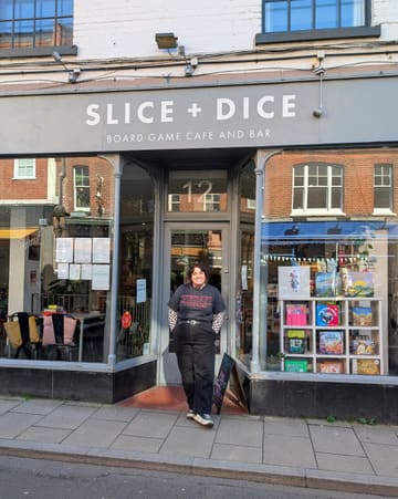 A photo of a creator standing in front of the 'Slice + Dice' board game cafe and bar in Norwich. The image serves as an introduction to a review of the venue, showing the storefront and the creator.