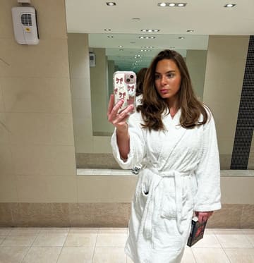A mirror selfie taken in a hotel or spa bathroom, featuring a woman wearing a white bathrobe.