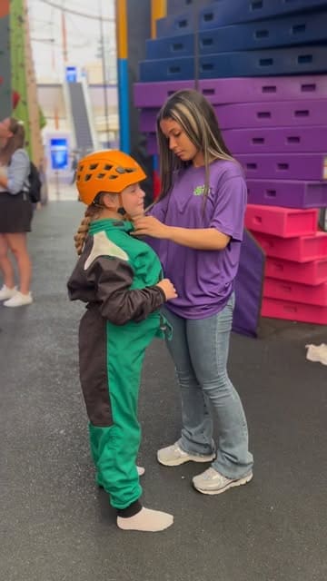 A clear, functional shot taken at Clip 'n Climb at The O2, showing a staff member helping a child put on a climbing harness. The background features the colourful climbing wall structure, establishing the venue's activity-focused environment.