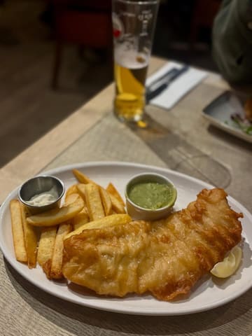 A standard, casual shot of a fish and chips meal at a gastropub. The image captures the meal and a drink in a typical pub setting, but lacks professional styling or careful framing.