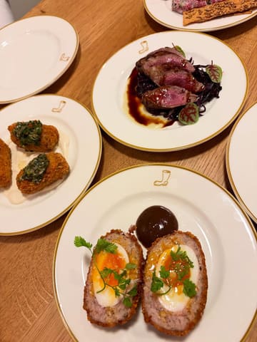 A flat-lay photograph of multiple small plates on a wooden table, featuring dishes like scotch eggs, venison, and beef tartare.
