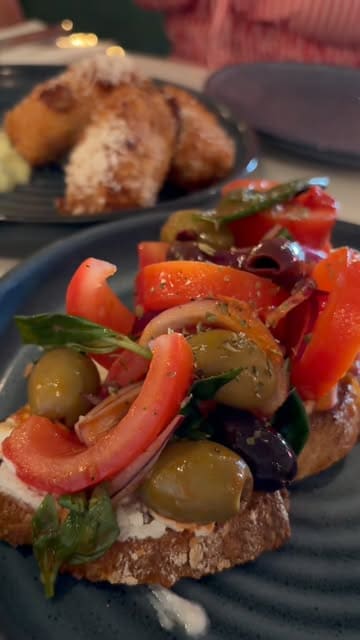 A close-up, appetizing shot of a bruschetta dish featuring fresh tomatoes, olives, and herbs, with a secondary plate of croquettes visible in the background, capturing a vibrant dining experience.