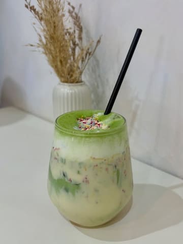 A clean, well-composed, and brightly lit shot of an iced matcha latte topped with sprinkles, presented in a glass against a minimalist white background with dried flowers.