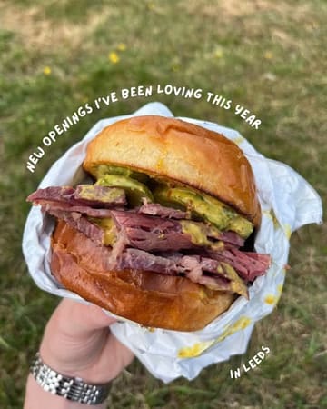 A close-up, handheld shot of a salt beef or pastrami sandwich in a paper wrapper, set against a grassy outdoor background, used to highlight a new food opening in Leeds.
