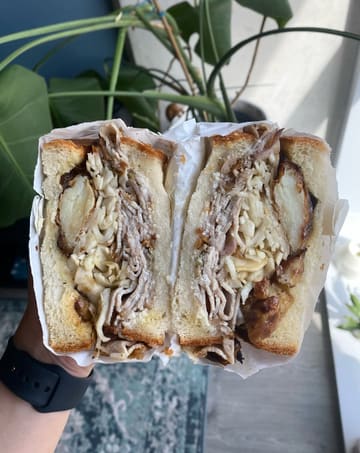 A high-quality, close-up shot of a porchetta sandwich held in hand, effectively showcasing the layers and textures of the ingredients. The natural lighting and shallow depth of field draw the viewer's eye directly to the cross-section of the sandwich.