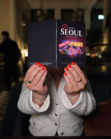 A person holds up a 'Seoul Food Tour Guide' menu from Six by Nico, obscuring their face. The image is taken in a dimly lit restaurant setting using a direct flash, which illuminates the menu clearly but leaves the background dark and creates a high-contrast look.