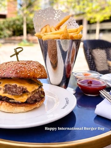 A close-up, eye-level shot of a double cheeseburger and a metal cone of fries served on a blue table, likely at an outdoor venue.