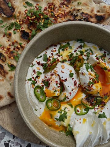 A high-quality, overhead food photography shot of Turkish eggs served with flatbread. The image captures the vibrant colors of the chili oil, fresh herbs, and the runny texture of the poached eggs, making for a highly appealing breakfast or brunch visual.