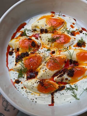 A vibrant, overhead shot of a homemade Turkish eggs dish, featuring a creamy yoghurt base topped with soft-boiled eggs, chili oil, and fresh dill, presented in a clean, minimalist bowl.
