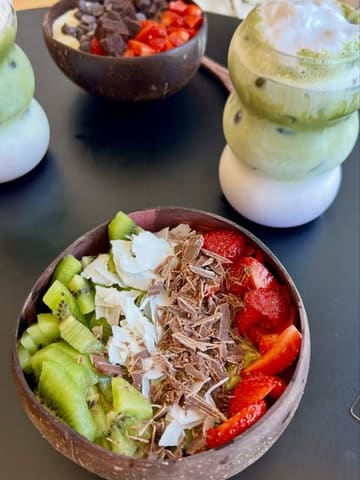 A top-down shot of two acai bowls and two layered iced matcha drinks on a black table. The image captures the vibrant colours of the fruit and the aesthetic presentation of the drinks, typical of a trendy cafe setting.