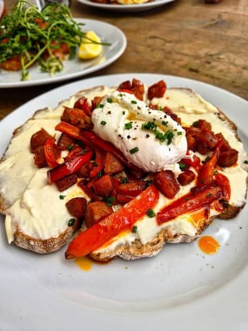 A high-quality, appetizing close-up shot of a brunch dish featuring chorizo, roasted red peppers, and whipped cream cheese on sourdough toast, topped with a poached egg. The lighting is natural and flattering, highlighting the textures of the food.