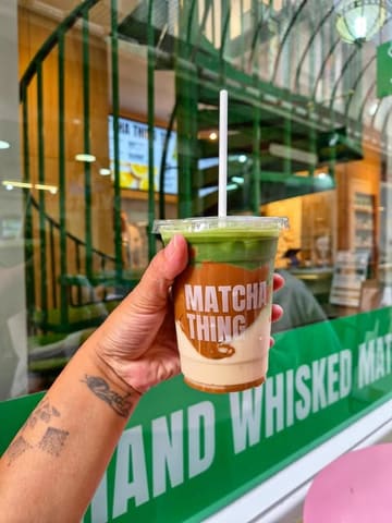A close-up shot of a hand holding a layered cookie butter matcha latte in front of a cafe window, showcasing the product branding.
