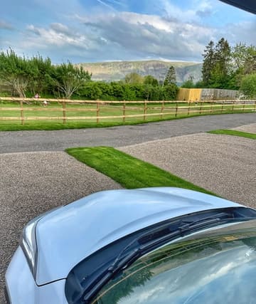 A scenic, point-of-view shot taken from inside a vehicle, showcasing a picturesque campsite in the Lake District with a stunning mountain backdrop. The image captures the peaceful, outdoor atmosphere of the location.