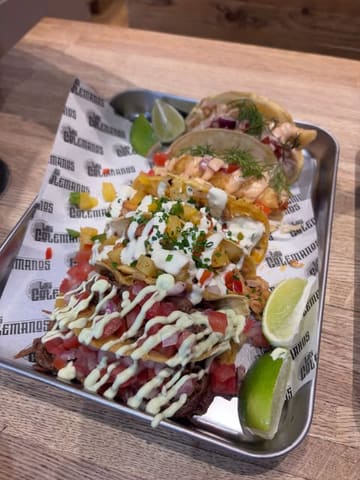A close-up, high-angle shot of a metal tray filled with various tacos, garnished with lime wedges and served on branded paper.