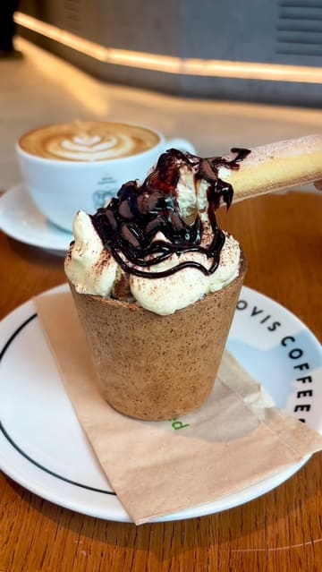 A short, aesthetic video showcasing a 'Cookie Cloud Cup' and a latte at Ovis Coffees. The video features close-up shots of the cookie cup being topped with cream and chocolate sauce, followed by a latte with latte art, and a spoon interacting with the dessert.