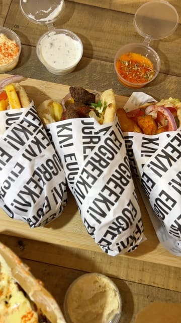 A handheld food haul video showcasing a variety of Greek takeaway dishes, including gyros, loaded fries, dips, and baklava, presented on a table for a group meal.