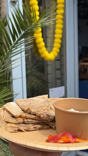 A short, energetic vlog-style video showcasing a small Indian street food stall called Tava Rakh in Peckham. The video features the exterior of the hut, the owner, and close-ups of various plant-based Indian dishes like samosa chaat and aloo paratha, capturing a casual and friendly atmosphere.