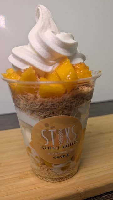 A close-up, satisfying video showing the assembly of a mango-based dessert in a cup, featuring layers of shredded pastry, soft-serve ice cream, and syrup.