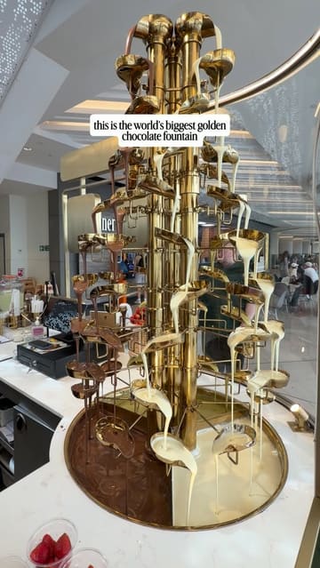 A promotional montage showcasing a dessert venue featuring a large golden chocolate fountain. The video highlights various desserts being prepared and drizzled with chocolate, including cheesecake, brownies, and fruit, set to music with text overlays.