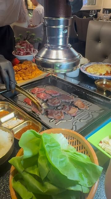 A top-down view of a Korean BBQ dining experience, showing raw meat being grilled on a table-top grill, accompanied by various side dishes and a basket of lettuce for wraps.