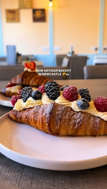A short, visually focused reel showcasing a new artisan bakery in Newcastle. The video features close-up shots of various pastries, including fruit-topped croissants, a tiramisu danish, and a blueberry brioche bun, alongside specialty coffees. It concludes with a shot of the bakery's exterior.