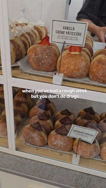 A short, handheld pan across a display case of various gourmet donuts at a cafe, accompanied by a relatable text overlay about using food as a coping mechanism.