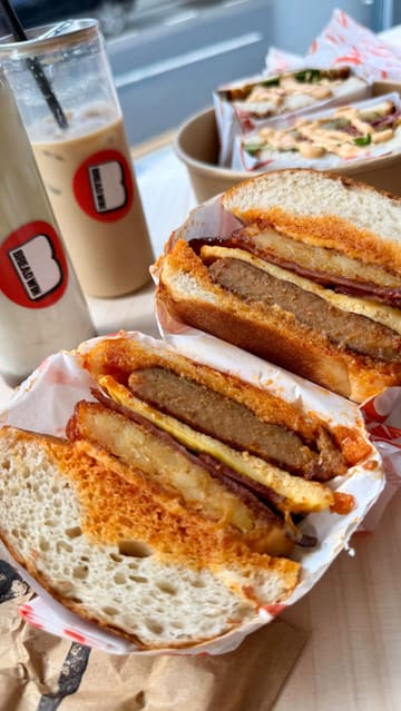 A short-form video review of a new sandwich shop in Newcastle called Breadwin. The creator showcases the interior, the ordering process, and several menu items including a breakfast bun, a pastrami sandwich, iced drinks, and pastries. The video uses a fast-paced, vlog-style format with voiceover and text overlays to highlight the food quality and venue atmosphere.