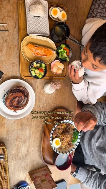 A video review of Mitsuba, a Japanese cafe in West London, featuring a mother and son trying various menu items including bento boxes, rice bowls, pastries, and cakes. The video highlights the cozy interior, specifically the tatami seating area, and provides a glimpse into the dining experience.