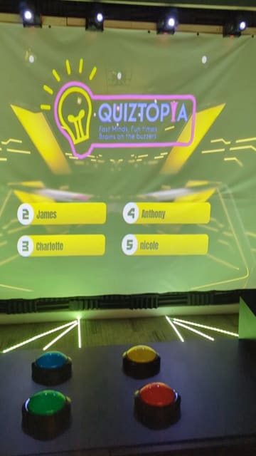 A promotional video for Quiztopia, an immersive quiz experience in Liverpool, featuring a group of friends playing the game, showing the buzzers, podiums, and game show-style screens.
