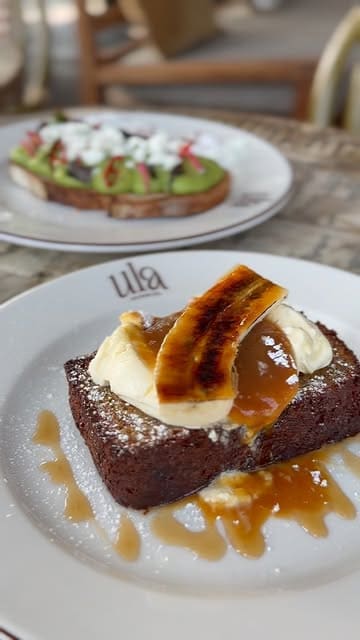 A high-quality, well-composed shot of a brunch spread featuring banana bread with cream and syrup in the foreground and avocado toast in the background. The lighting is soft and natural, highlighting the textures of the food effectively.