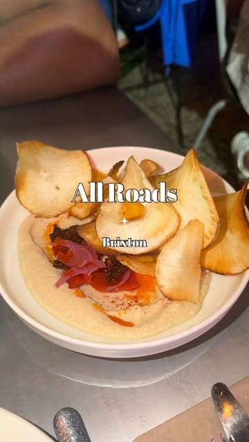 A review of All Roads in Brixton, showcasing a variety of dishes including cassava chips, lamb ribs, fried wings, turkey skewers, mac and cheese, cornbread, and peach cobbler, while highlighting the lively atmosphere with a DJ.