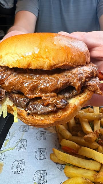 A creator reviews a burger restaurant in Manchester, showcasing several menu items including smash burgers with brisket, chicken tenders, and loaded fries. The video also highlights the venue's 'bunker' games room and film club, positioning it as a great spot for parties and social gatherings.