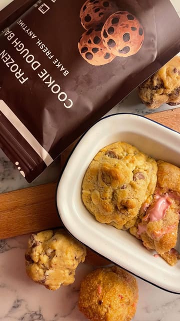 A short, handheld video demonstrating the process of baking frozen cookie dough balls from BIY Cookies. The creator shows the packaging, places the frozen dough into a dish, and reveals the final baked, gooey cookies.