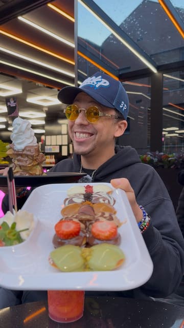 A high-energy, creator-led review of Rassam's Creamery, a dessert venue in Liverpool. The video highlights the futuristic interior, the use of robot waiters, and the variety of indulgent desserts like waffles and mini Dutch pancakes. It also includes a cross-promotion tip for a discount when visiting a nearby burger restaurant.