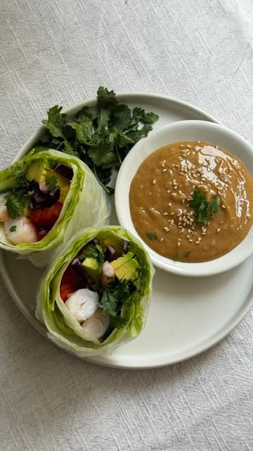 A clean, top-down photograph of homemade summer rolls served with a peanut dipping sauce. The rolls are filled with prawns, avocado, peppers, and cabbage, presented on a simple white plate against a textured white background, showcasing fresh and vibrant ingredients.