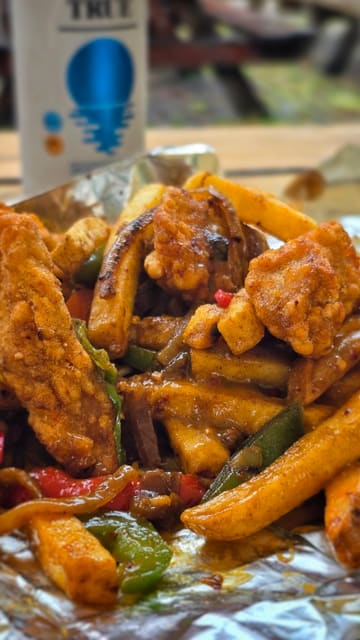 A short, handheld video showcasing a 'spice bag' takeaway meal from a food truck. The creator opens the branded paper bag to reveal the contents, including chicken strips, seasoned fries, peppers, onions, and a side of curry sauce, paired with a non-alcoholic lager.