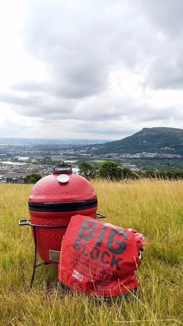 A man prepares a breakfast burrito on a portable Kamado Joe grill while on a hill overlooking Belfast. The video follows the cooking process from lighting the charcoal to assembling the burrito with bacon, sausage, egg, relish, and avocado.