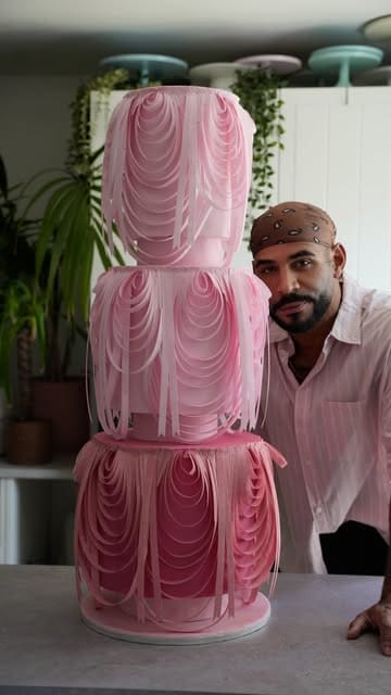 A creator demonstrates the assembly and decoration of a large, tiered pink cake using wafer paper techniques in a studio setting.