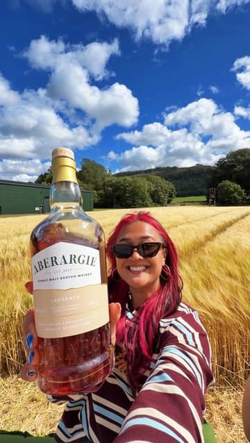A vibrant vlog-style video documenting a group visit to Aberargie Distillery in Scotland. The video features a tour of the production facilities, including the stills and barrel warehouse, followed by an outdoor tasting session and meal in a picturesque barley field. The creator highlights two new whisky expressions, 'Laureate' and 'Golden Promise', while showcasing the social and educational aspects of the distillery experience.