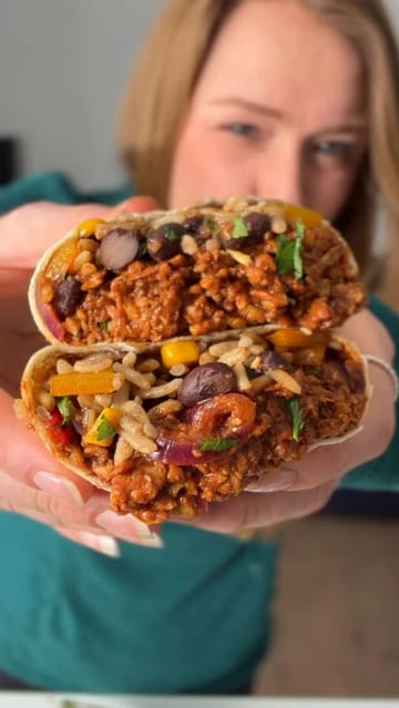 A short, instructional video demonstrating how to assemble a vegan tempeh wrap. The creator shows the layering of ingredients including rice, corn, beans, and seasoned tempeh, finishing with a bite of the final product.