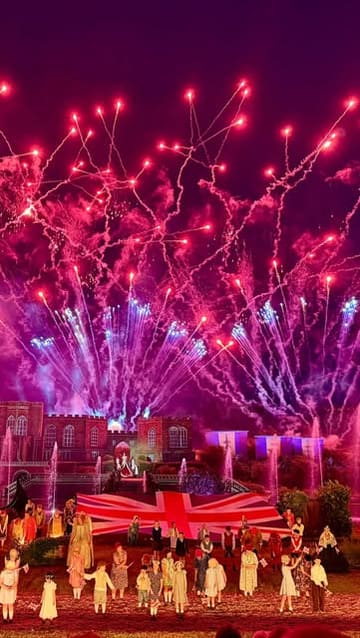 A wide-angle, night-time shot of the Kynren live-action show, capturing a large Union Jack flag on stage, performers, and a dramatic fireworks display illuminating the sky.