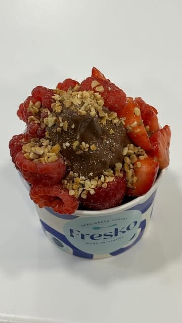 A short, visually appealing video showcasing the assembly of a frozen yogurt dessert with chocolate sauce, fresh strawberries, raspberries, and crushed nuts at a Fresko venue.