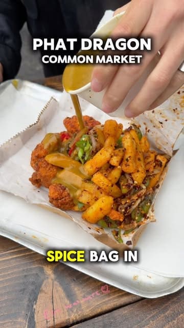 A close-up, top-down action shot of a spice bag being drizzled with curry sauce, highlighting the crispy chicken, chips, and peppers in a takeaway tray.