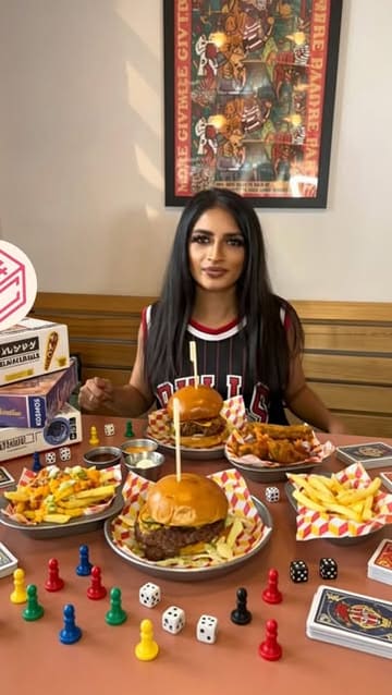 A promotional lifestyle shot featuring a person at a table in a board game cafe, surrounded by burgers, fries, and various game components, effectively showcasing the venue's unique selling point.