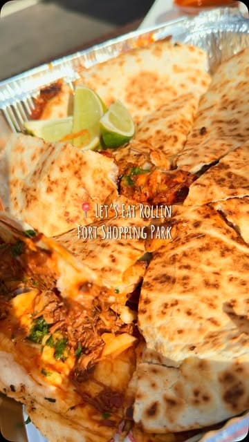 A short promotional video for a food truck called Let's Eat Rollin located at Fort Shopping Park, showcasing their birria quesadilla, pizza, and tacos served with consommé.