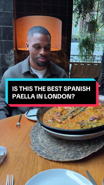 A food review video featuring a visit to Arros QD in London. The creator samples various dishes including paella, lamb short ribs, carabineros prawn, king scallop, and basque cheesecake, while also highlighting the open-fire cooking process in the kitchen.