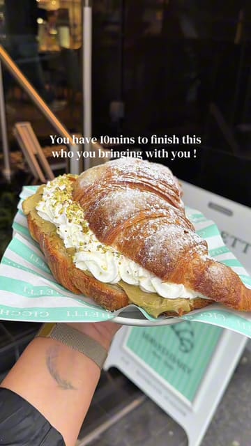 The video features a close-up look at a giant croissant being prepared and served at San Carlo Cicchetti, alongside other breakfast items like eggs benedict and a yogurt bowl. The footage captures the preparation process and the final presentation in the restaurant setting.